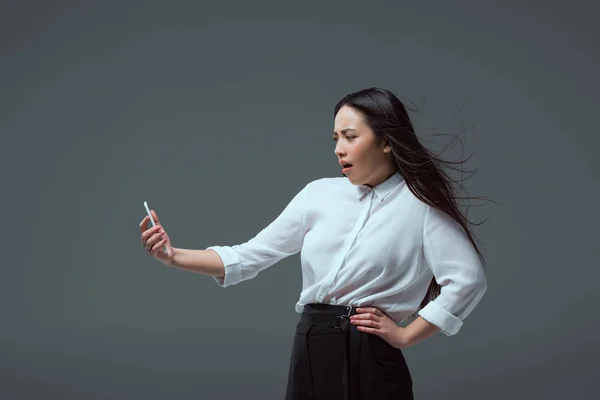 Shocked young asian woman holding smartphone isolated on grey — Stock Photo