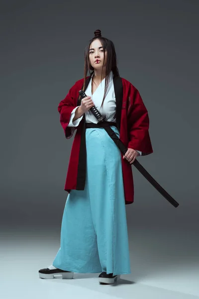 Samurai en kimono sosteniendo espada katana y mirando a la cámara en gris - foto de stock