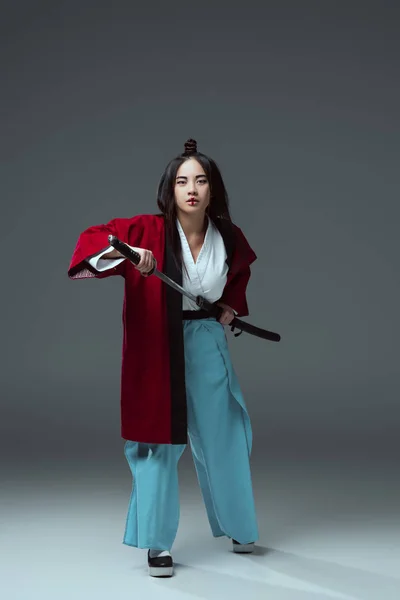 Full length view of young asian woman in kimono holding katana sword and looking at câmera on grey — Fotografia de Stock