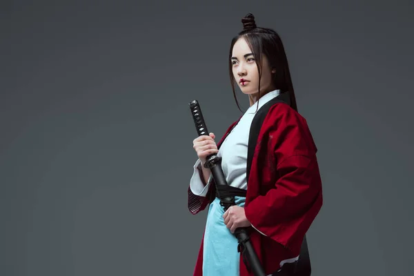 Joven asiático mujer en kimono holding katana y mirando lejos aislado en gris - foto de stock