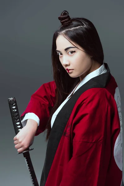 Молодая азиатка в кимоно держит катану и смотрит на камеру, изолированную на сером — стоковое фото