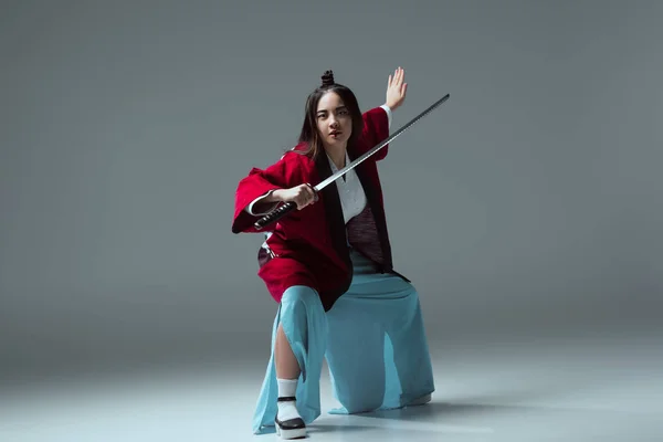 Mujer asiática en kimono luchando con espada katana y mirando a la cámara en gris - foto de stock