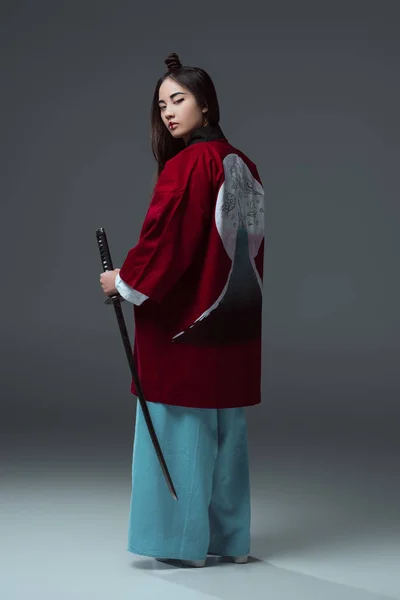 Вид сзади женщины в традиционном кимоно, держащей Фанану и смотрящей в камеру на сером — стоковое фото