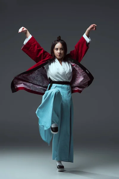 Повний вигляд на азіатську жінку в традиційному японському кімоно, дивлячись на камеру на сірому — стокове фото