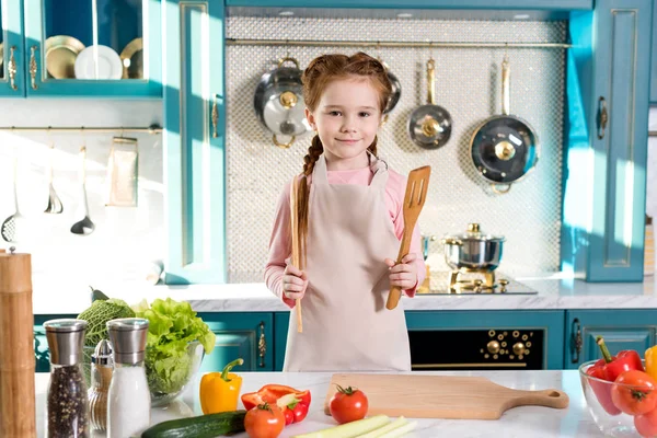 Очаровательный ребенок в фартуке держа деревянную посуду и улыбаясь на камеру на кухне — стоковое фото