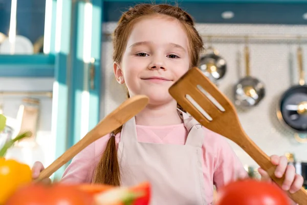 Beautiful happy child in apron holding wooden utensils and smiling at camera — Stock Photo