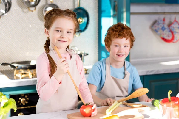 Entzückend glückliche Kinder in Schürzen lächeln in die Kamera, während sie gemeinsam in der Küche kochen — Stockfoto