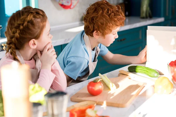 Діти в фартухах читають кубок під час приготування овочевого салату разом на кухні — стокове фото