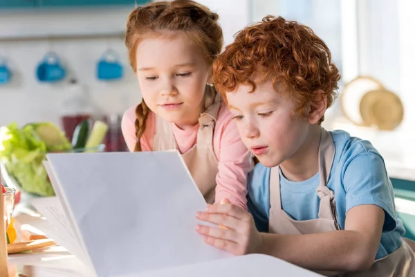 Niños pequeños enfocados leyendo el libro de cocina mientras cocinan juntos en la cocina - foto de stock