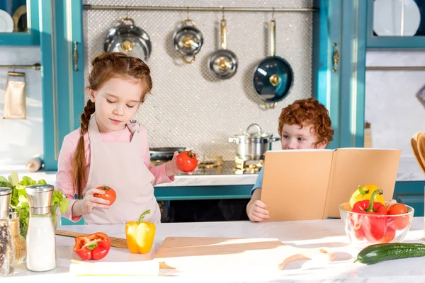 Niños con libro de cocina y verduras cocinando juntos en la cocina - foto de stock