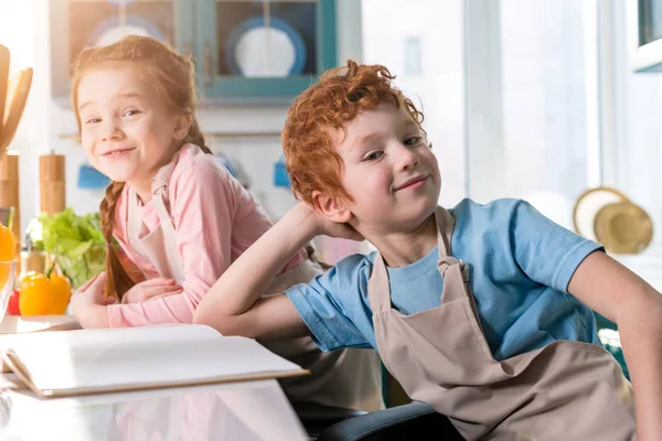 Adorables enfants dans des tabliers souriant à la caméra tout en cuisinant avec livre de cuisine dans la cuisine — Photo de stock