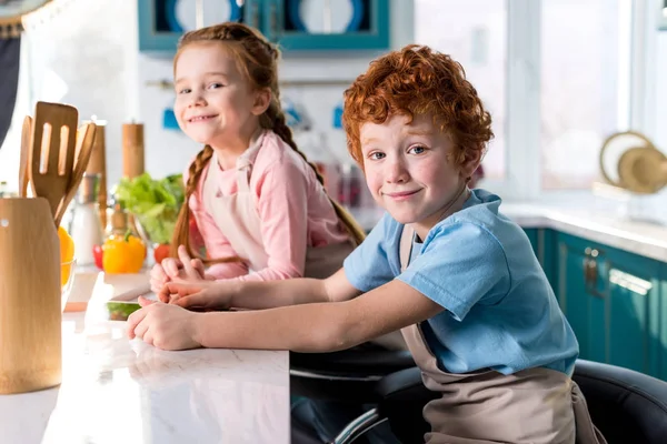 Очаровательные счастливые дети улыбаются в камеру во время приготовления пищи вместе на кухне — стоковое фото