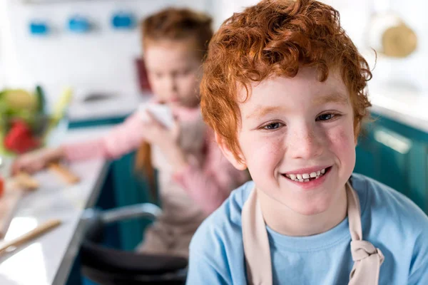 Carino bambino sorridente a macchina fotografica mentre amico cucina dietro in cucina — Foto stock