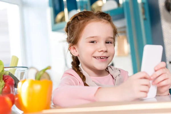 Adorable child smiling at camera while using smartphone in kitchen — Stock Photo