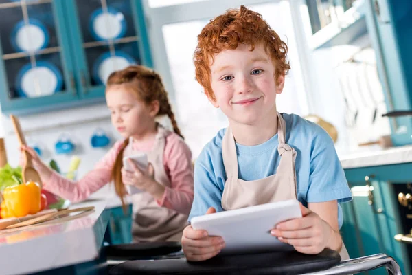 Netter kleiner Junge mit digitalem Tablet lächelt in die Kamera, während das Kind mit dem Smartphone im Hintergrund kocht — Stockfoto