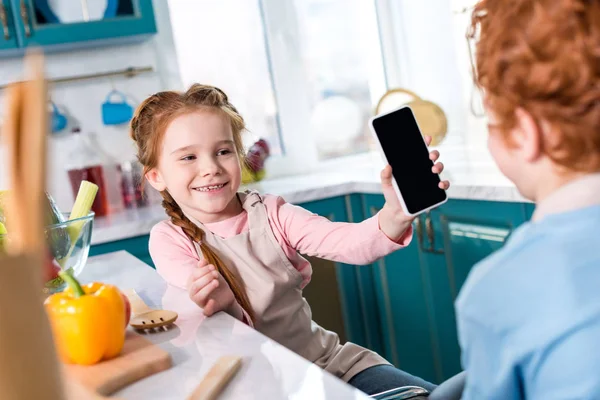 Recortado de los niños usando el teléfono inteligente con pantalla en blanco mientras cocinan juntos en la cocina - foto de stock