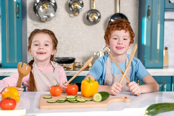 Entzückende Kinder, die Holzutensilien in der Hand halten und beim gemeinsamen Kochen in der Küche in die Kamera lächeln — Stockfoto