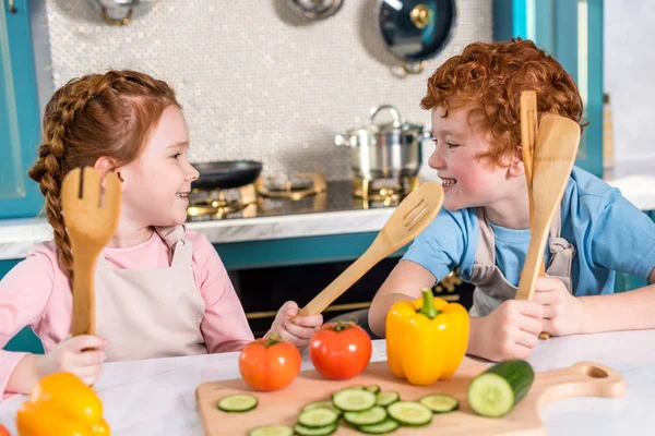 Crianças felizes com utensílios de madeira sorrindo uns aos outros enquanto cozinham juntos na cozinha — Fotografia de Stock