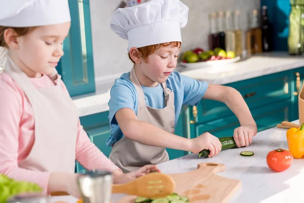 Enfants dans des chapeaux de chef et des tabliers préparant ensemble une salade de légumes dans la cuisine — Photo de stock