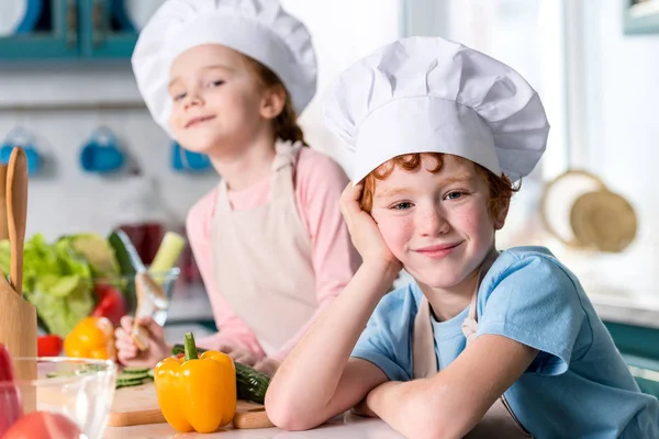 Adorable siblings in chef hats and aprons smiling at camera while cooking in kitchen — Stock Photo