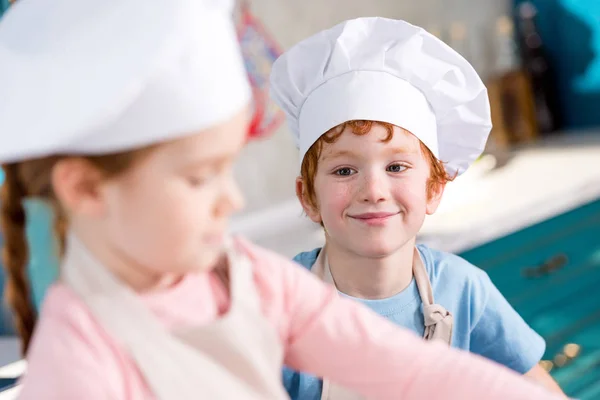 Adorable smiling little boy in chef hat looking at friend cooking on foreground — Stock Photo