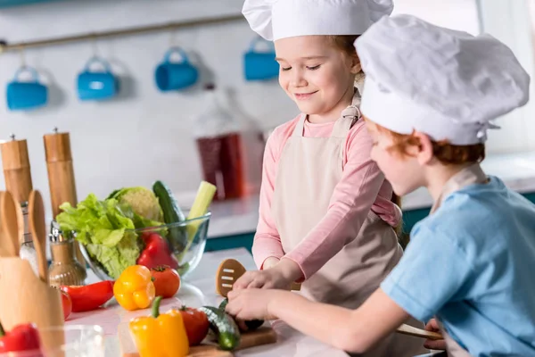 Niedliche kleine Kinder in Kochmützen bereiten gemeinsam in der Küche Gemüsesalat zu — Stockfoto