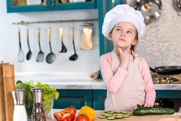 Niño reflexivo en sombrero de chef y delantal mirando hacia otro lado en la cocina - foto de stock
