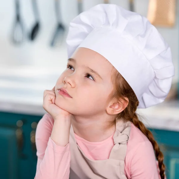 Cher petit enfant au chapeau de chef tenant la main sur le menton et regardant loin dans la cuisine — Photo de stock