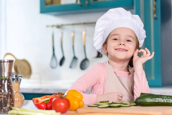 Очаровательный ребенок в шляпе шеф-повара показывая ОК знак и улыбаясь на камеру на кухне — стоковое фото