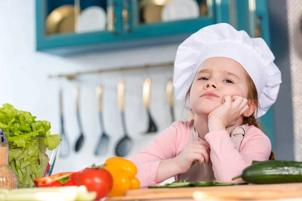Enfant ennuyé dans le chapeau de chef assis avec la main sur le menton dans la cuisine — Photo de stock