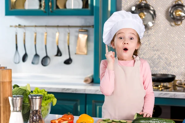 Niño emocionado en sombrero de chef y delantal apuntando hacia arriba con el dedo y mirando a la cámara en la cocina - foto de stock