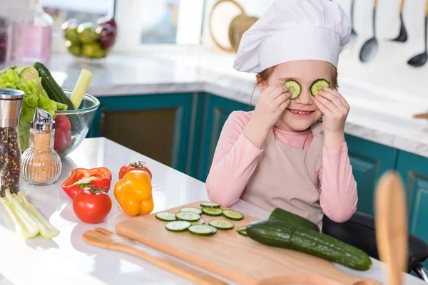 Criança sorridente em chapéu de chef e avental segurando fatias de pepino enquanto cozinha na cozinha — Fotografia de Stock