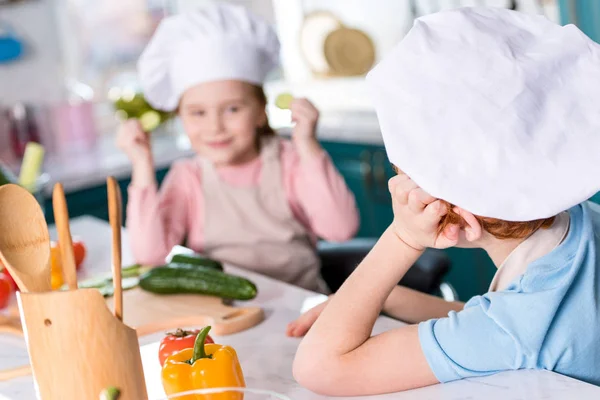 Очаровательные дети в шляпах шеф-повара глядя друг на друга во время приготовления пищи вместе на кухне — стоковое фото