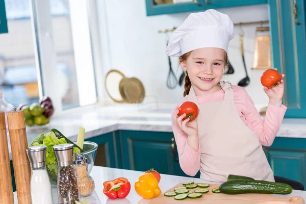 Nettes Kind mit Kochmütze und Schürze, das Tomaten hält und in die Kamera lächelt, während es in der Küche kocht — Stockfoto