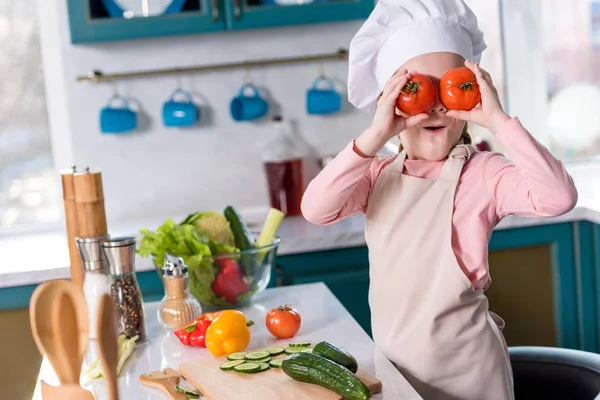 Cute child in chef hat and apron holding tomatoes while cooking in kitchen — Stock Photo