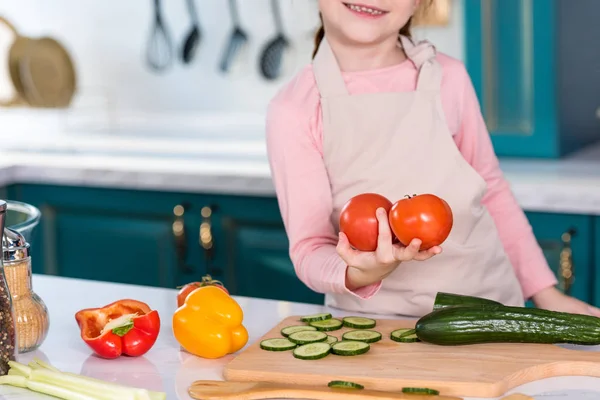 Обрезанный снимок улыбающегося ребенка в фартуке, держащего свежие помидоры на кухне — стоковое фото