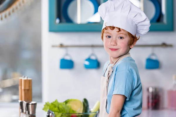 Lindo niño en sombrero de chef y delantal sonriendo a la cámara en la cocina - foto de stock
