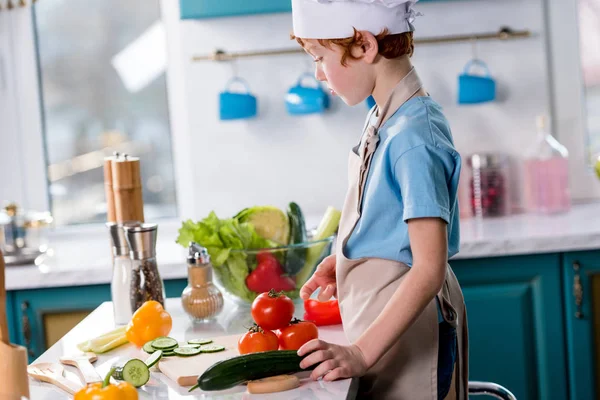 Vista lateral de lindo niño en sombrero de chef y delantal de cocina en la cocina - foto de stock