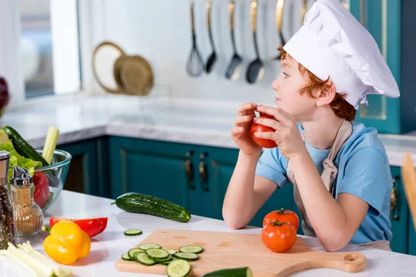Lindo niño en sombrero de chef y delantal mirando hacia otro lado mientras está sentado en la cocina - foto de stock