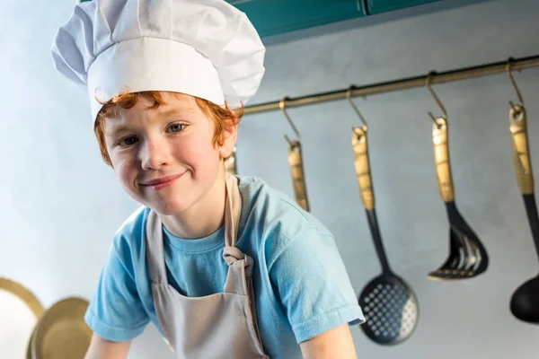 Очаровательный маленький мальчик в шляпе шеф-повара и фартуке, улыбающийся в камеру на кухне — стоковое фото