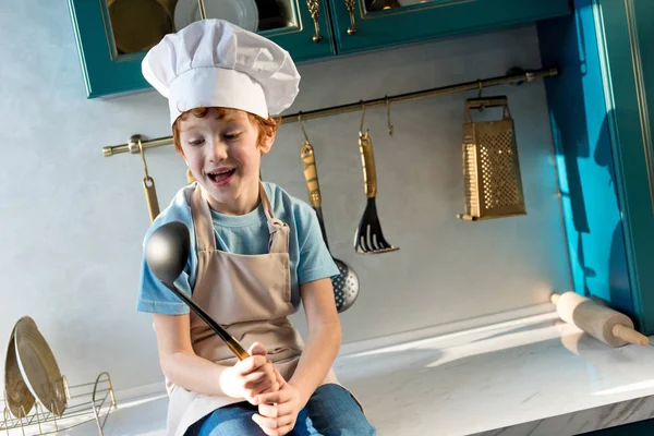 Mignon petit garçon heureux dans le chapeau de chef et tablier tenant louche dans la cuisine — Photo de stock