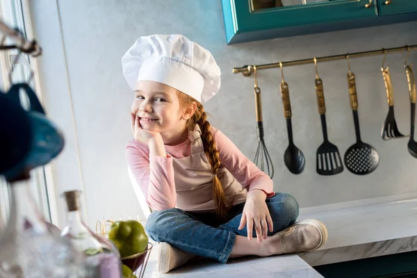 Criança adorável em chapéu de chef sentado na cozinha e sorrindo para a câmera — Fotografia de Stock
