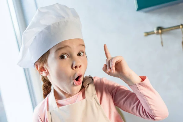 Nettes Kind mit Kochmütze, das mit dem Finger nach oben zeigt und in der Küche in die Kamera schaut — Stockfoto