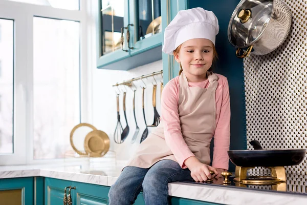 Criança sorridente bonito em chapéu de chef e avental olhando para frigideira no fogão — Fotografia de Stock