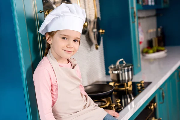 Adorable niño en sombrero de chef y delantal sonriendo a la cámara en la cocina - foto de stock