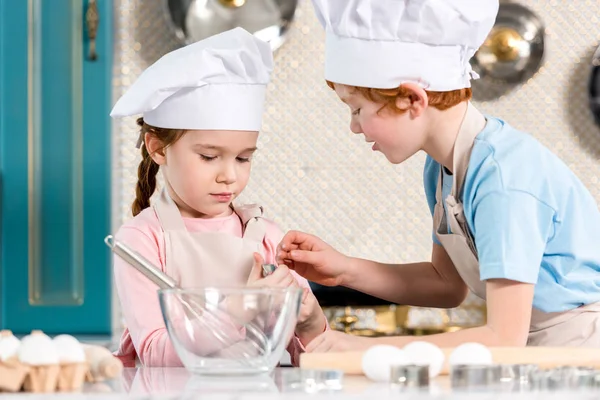 Красиві маленькі діти в капелюхах шеф-кухаря і фартухах, які готують разом на кухні — стокове фото