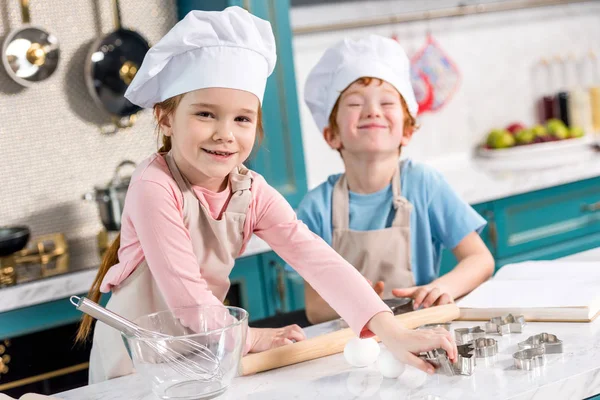 Очаровательные маленькие дети в шляпах и фартуках шеф-повара, улыбающиеся в камеру, пока готовят вместе на кухне — стоковое фото