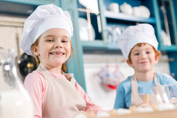 Crianças adoráveis em chapéus de chef e aventais sorrindo para a câmera enquanto cozinham juntos na cozinha — Fotografia de Stock