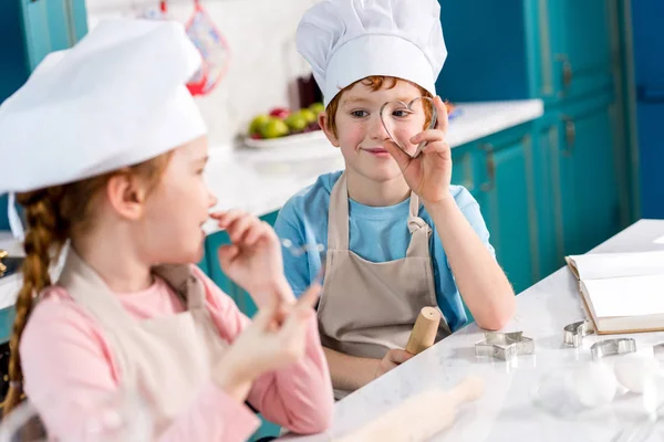 Entzückende Kinder in Kochmützen und Schürzen, die einander beim gemeinsamen Kochen in der Küche lächelnd begegnen — Stockfoto