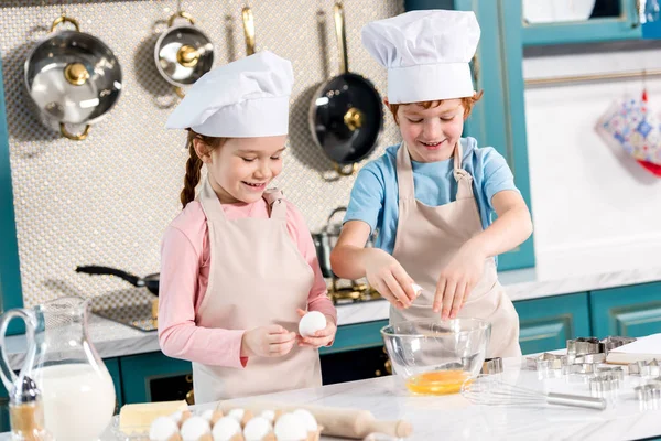 Милые улыбающиеся дети в шляпах шеф-повара и фартуках готовят тесто вместе на кухне — стоковое фото